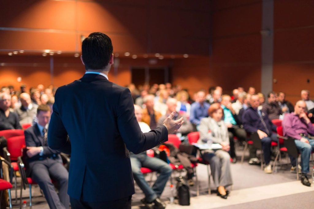 Image of back of man standing in front of a room of seated people speaking at a trade show event